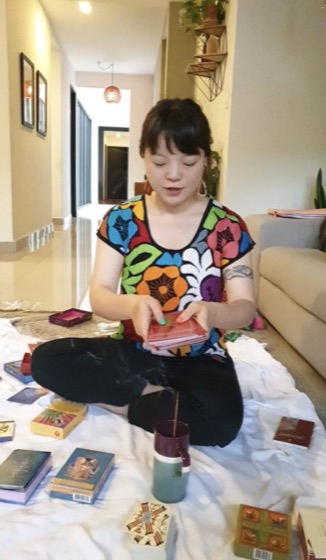 Simone sitting on the floor in a colorful floral top, shuffling a tarot deck surrounded by oracle card boxes and incense