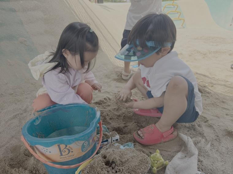 Two toddlers digging in the sand together with a blue bucket