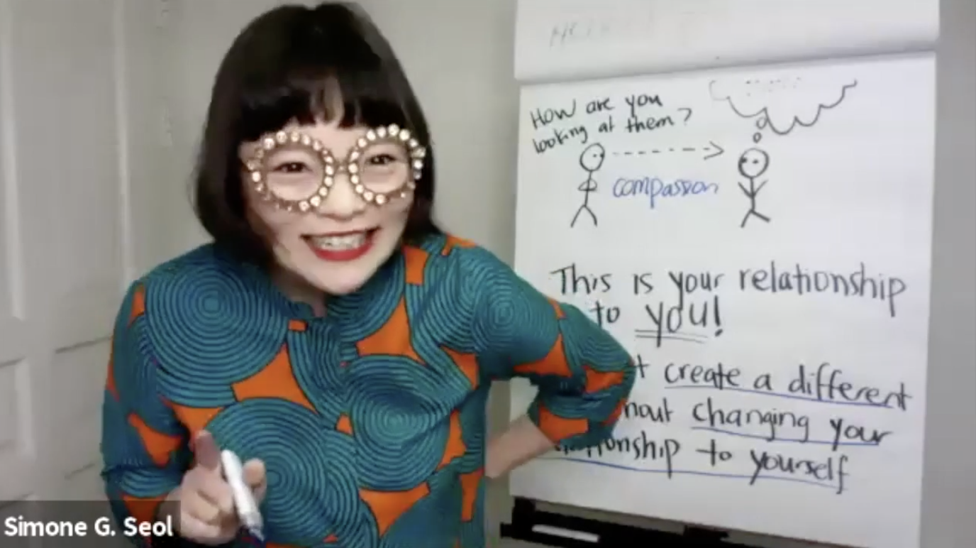 Simone Grace Seol teaching at the whiteboard, wearing bedazzled glasses, gesturing animatedly during the Model Heresy class.